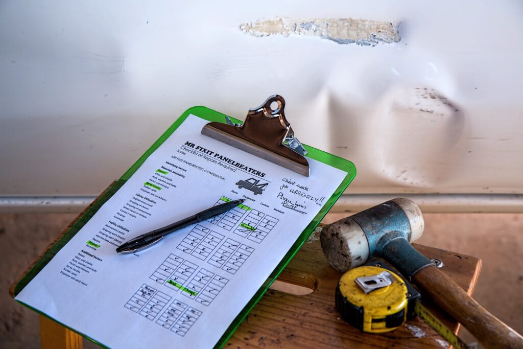 A green clipboard holding a 'Mr Fixit Panelbeaters' repair checklist with a pen, resting on a wooden stool beside a rubber mallet and yellow tape measure against a dented white panel.