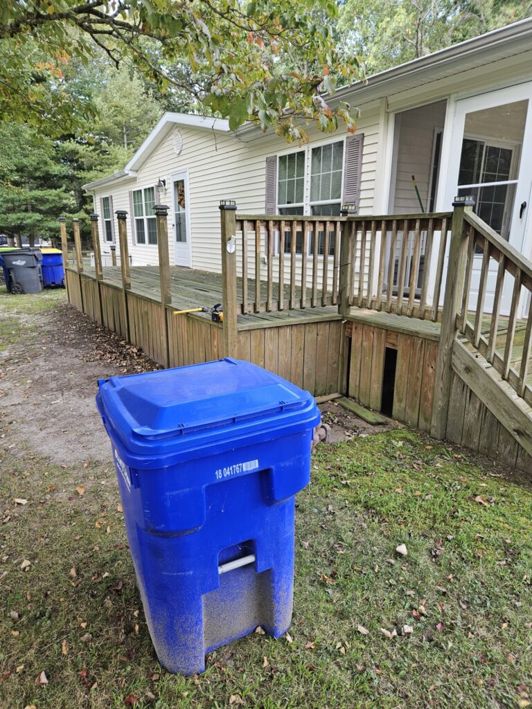 A blue recycling bin sits on grass in a backyard next to a wooden deck attached to a cream-colored house, with trees in the background.