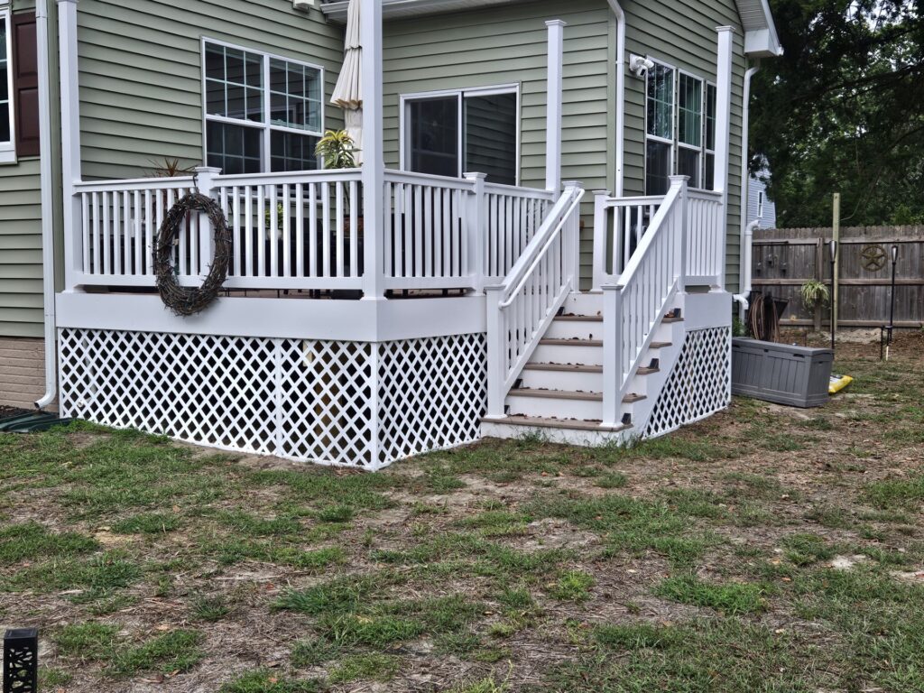 Raised back deck with white vinyl railings, lattice skirting, and stairs with brown treads beside a sage green house