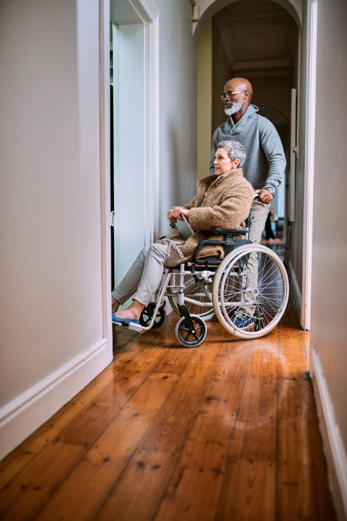 Older man pushes a woman in a wheelchair down a hallway with wide plank wood floors, white baseboards and an arched doorway