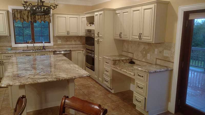 Kitchen with cream raised-panel cabinets, granite island, built-in desk, double wall oven and travertine tile backsplash