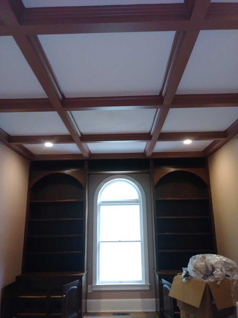 Stained wood coffered ceiling above dark built-in arched bookcases flanking an arched window, with recessed lights
