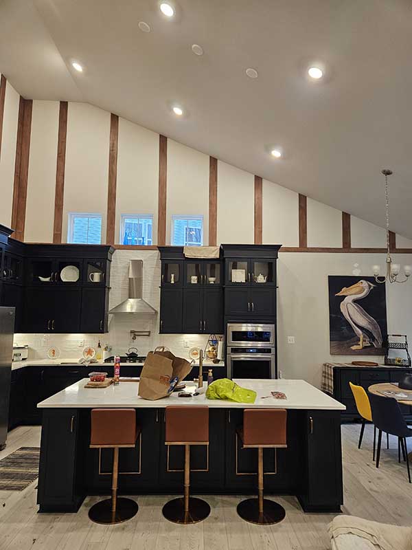 Kitchen with black cabinets, white island, three leather barstools, and vaulted ceiling with stained wood trim strips