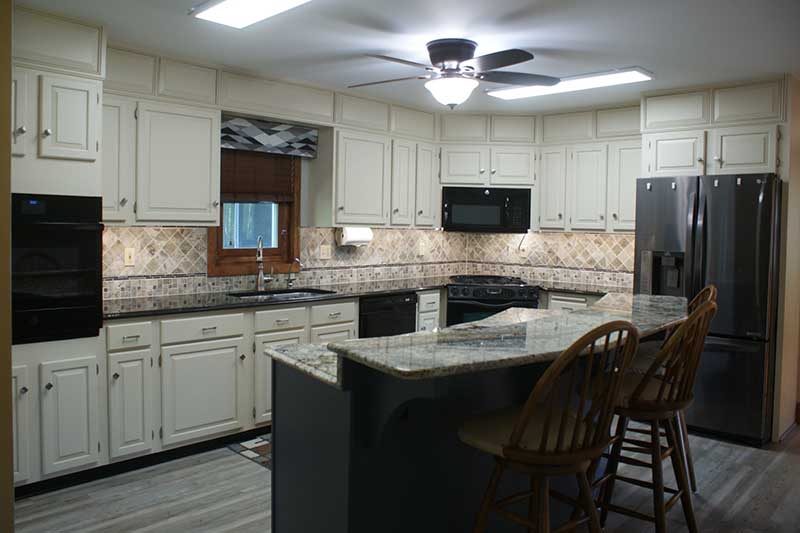 Kitchen with cream raised-panel cabinets, granite island with wood stools, tile backsplash, black appliances, gray plank floor