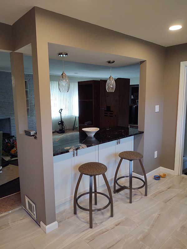 Kitchen pass-through with dark countertop, white base cabinets, two metal bar stools, pendant lights, and light plank flooring