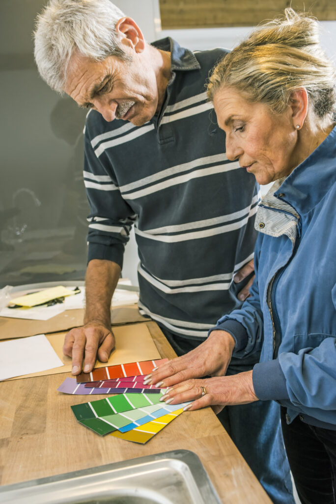 Older man and woman studying fanned paint color swatch cards in red, purple, green, blue, and yellow on a wooden countertop