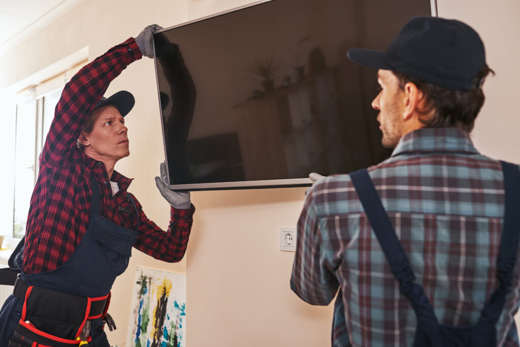 Two technicians in plaid shirts, caps and overalls lift a flat-screen TV onto a beige living room wall