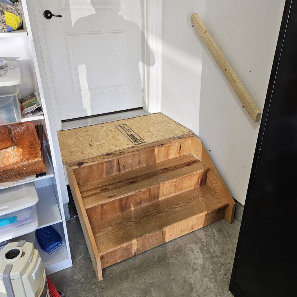 Custom wooden steps with OSB landing leading to a white interior door in a garage, with a wood handrail on the wall