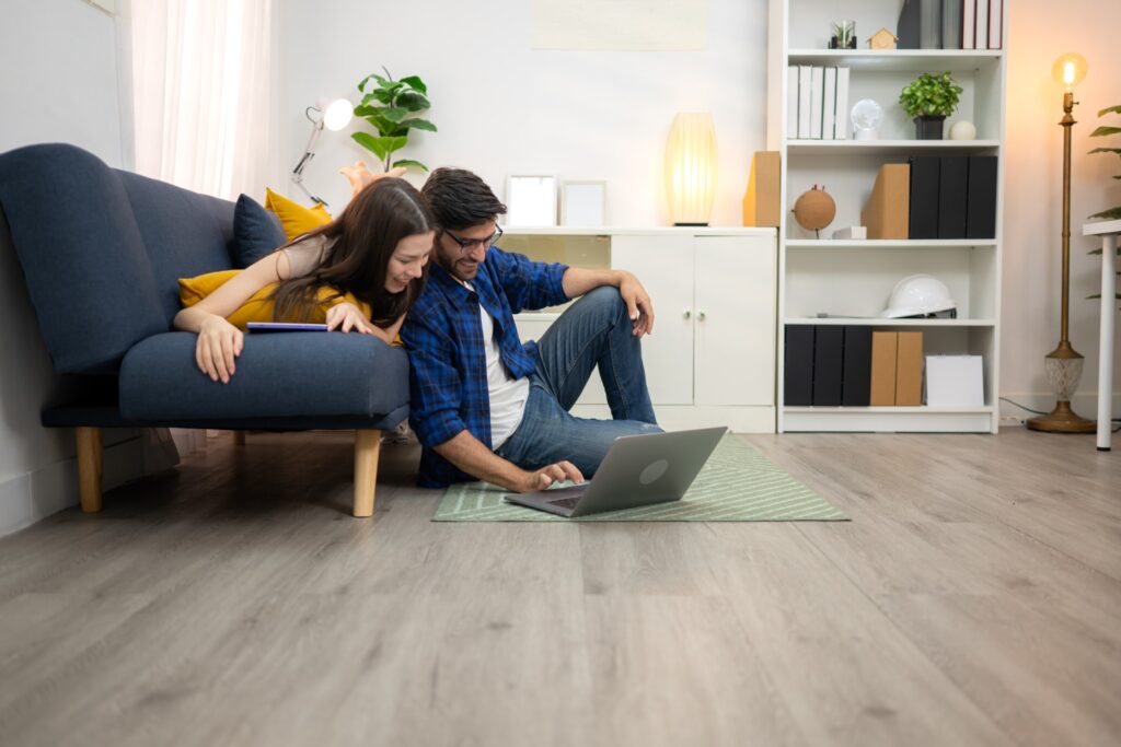 Couple using a laptop in a living room with light gray wood-look plank flooring, blue sofa, and white bookshelf