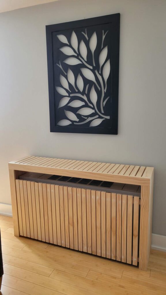 Unfinished pine radiator cover with vertical slats and slatted top below a dark leaf-pattern wall panel on a gray wall