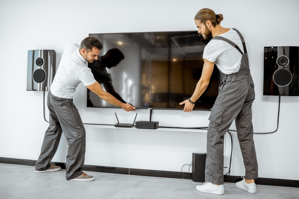 Two workers in gray work pants positioning a large flat-screen TV on a white wall between two wall-mounted speakers